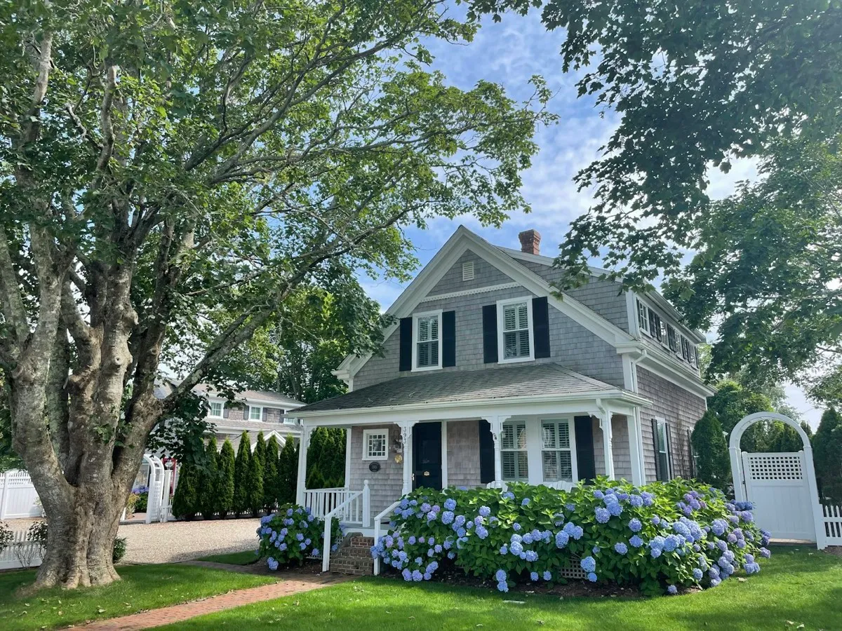 Gray wooden house with green trees and garden beds